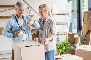 Woman in striped shirt and man in blue shirt unpacking brown box, surrounded by unopened boxes in new apartment