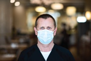 Man with short hair and black shirt facing camera wearing blue medical mask